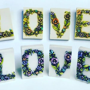 May include: A set of eight ceramic tiles spelling out the word "LOVE" in all capital letters. Each letter is surrounded by a wreath of colorful flowers.