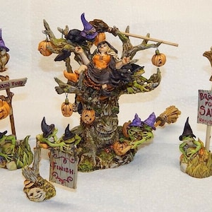 May include: A Halloween-themed figurine set featuring a witch sitting on a tree with a crow and pumpkins. There are also frogs wearing hats and holding brooms, and signs that say "Lesson Fund" and "Brooms for Sale".
