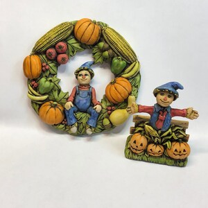 Puede incluir: Dos figuritas de cerámica de un espantapájaros y un niño, ambos con sombreros azules y monos. El espantapájaros está sentado en un banco con calabazas y maíz. El niño está sentado en una corona hecha de calabazas, maíz y otras verduras de otoño.