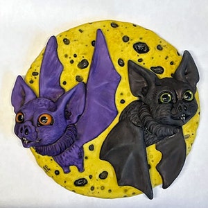 May include: Two ceramic bats with painted details, one purple and one black, are perched on a yellow moon-shaped background with black spots.