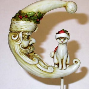 May include: A white ceramic ornament shaped like a crescent moon with a Santa Claus face and a white cat wearing a red Santa hat sitting on the moon.