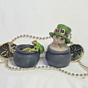 Può includere: Due piccoli vasi neri con monete d'oro all'interno. Un vaso ha una rana verde seduta sul bordo e l'altro ha un gatto bianco con un cappello da folletto verde.