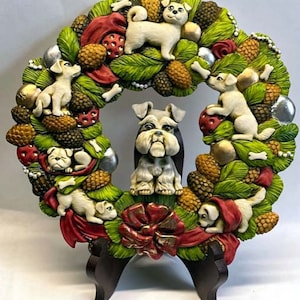 Puede incluir: Una corona de Navidad hecha de hojas verdes, piñas y lazos rojos. Hay varios perros pequeños blancos y un perro gris más grande en el centro de la corona.