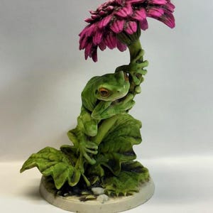 May include: A detailed figurine of a green frog perched on a flower stem, beneath a large, vibrant pink flower. The frog has orange eyes and is surrounded by green leaves. The base is a light beige color.
