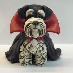 May include: A ceramic figurine of a Schnauzer dog dressed in a black cape and a red collar. The dog has a white beard and eyebrows, with black fur on its head and back. The cape is red and black, giving the dog a vampire-like appearance.