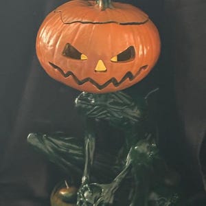 May include: A green ceramic figurine of a skeletal figure with a jack-o'-lantern head. The figurine is sitting with its hands clasped together and has a small pumpkin at its feet.