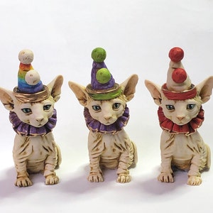 May include: Three ceramic Sphynx cat figurines, each wearing a different colored clown hat and ruffled collar. The hats are adorned with gold trim and pom-poms. The cats have pale skin and blue eyes, with a whimsical and decorative design.