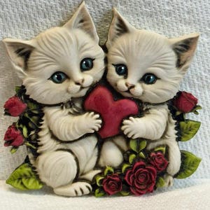 May include: A decorative figurine featuring two white kittens with blue eyes, holding a red heart. The kittens are surrounded by red roses and green leaves. The figurine has a vintage, romantic aesthetic.