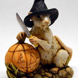 May include: A small figurine of a light-colored squirrel wearing a black pointed witch hat. The squirrel holds a silver-colored knife with a yellow handle and rests its hand on a carved orange pumpkin. The base is textured like brown stones.