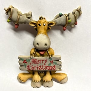 以下が含まれることがあります： 茶色と白の体をしたセラミック製のヘラジカの置物で、木製の看板を持っており、「メリークリスマス」と書かれています。ヘラジカは胸に赤いハートがあり、角は金色とカラフルなライトで飾られています。