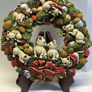 May include: A white ceramic Christmas wreath with a red bow and white kittens. The wreath is decorated with pine branches, pine cones, and silver ornaments. The wreath is on a brown wooden stand.