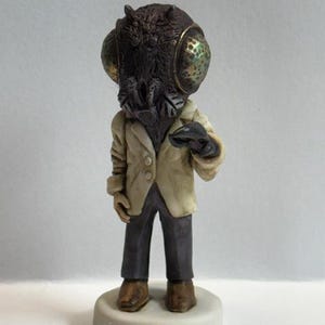 May include: A figurine of a person with a fly head wearing a white jacket and dark pants. The fly head has large, green eyes and a brown body.
