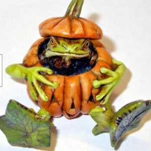 Puede incluir: Una figurita de rana verde que lleva un disfraz de calabaza naranja tallada. La calabaza tiene una cara de calabaza tallada y la rana sostiene la calabaza con sus manos.