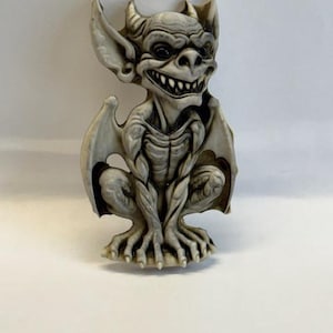 May include: A detailed, off-white gargoyle figurine with a menacing expression. The creature has large, bat-like wings, horns, and sharp teeth. It is in a crouching position, with its hands resting on its knees. The figurine is a decorative item.