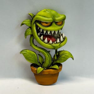 May include: A sculpted figure of a green carnivorous plant with a wide, toothy mouth, orange eyes, and a red tongue, emerging from a small brown terracotta-style pot. The plant features sharp white teeth and a dripping green substance.