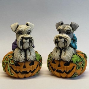 May include: Two white miniature schnauzer dog figurines sitting on top of orange jack-o'-lantern pumpkins with green vines. The dog on the left is wearing a purple bow and the dog on the right is wearing a blue bow.