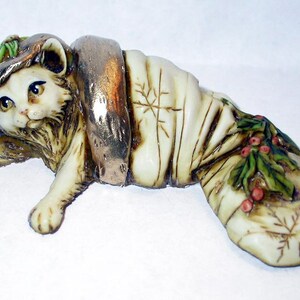 Könnte beinhalten: Eine weiße Keramikfigur eines Katers, der eine Strumpfkappe mit Stechpalme und Beeren trägt. Der Kater liegt auf der Seite, der Kopf ruht auf dem Strumpf.