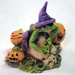 Op de afbeelding: Een groene kikker met een paarse heksenhoed zit op een bed van groene bladeren en houdt een kleine pompoen vast. Twee andere pompoenen bevinden zich naast de kikker, een met een gesneden gezicht en de andere met een eenvoudig pompoengezicht.