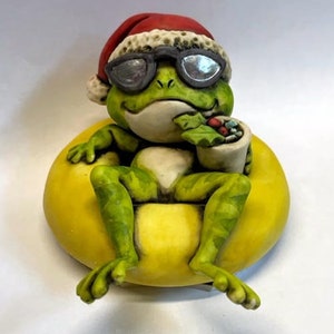 May include: A green frog wearing a red Santa hat and sunglasses sits in a yellow inflatable ring, holding a cup of red and green candies.