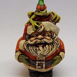 Può includere: Una stravagante statuetta natalizia di Babbo Natale con un volto sorridente, che indossa un abito rosso e una cintura nera. Una rana verde con un cappello è appollaiata sulla testa di Babbo Natale. Una decorazione festiva.