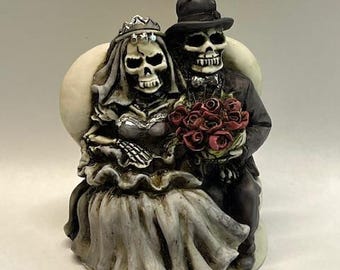 May include: A skeletal bride and groom wedding cake topper. The bride is wearing a white gown with a veil and a tiara. The groom is wearing a black suit and a top hat. They are holding a bouquet of red roses.