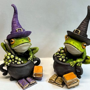 May include: Two green frog figurines wearing witch hats and holding cauldrons filled with green grapes. Each frog is sitting on a stack of books.