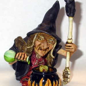 May include: A witch figurine with a black hat and long white hair, stirring a cauldron with a bone-handled broom. A black cat sits on top of the broom handle. The cauldron is on a base with flames.