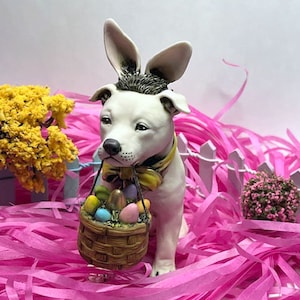 May include: A white ceramic dog figurine wearing bunny ears and a yellow bow tie, holding a basket of colorful Easter eggs. The figurine is surrounded by pink shredded paper.