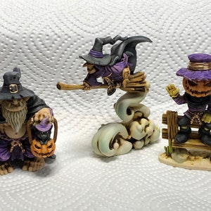 May include: Three Halloween themed figurines. A purple and black witch riding a broomstick, a purple and brown goblin holding a pumpkin, and a purple and brown jack-o-lantern holding a pumpkin.
