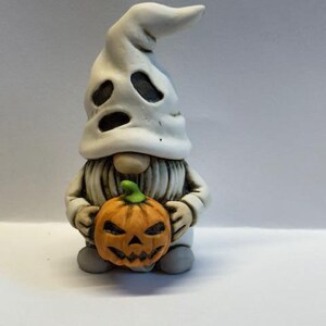 Puede incluir: Un gnomo de cerámica blanco que lleva un disfraz de fantasma y sostiene una calabaza de Halloween.