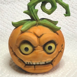 Peut inclure: Une figurine de citrouille en céramique avec une tige verte et une expression faciale menaçante. La citrouille a un large sourire aux dents, de grands yeux et une surface texturée.