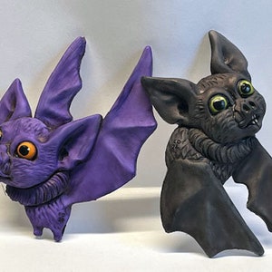 May include: Two ceramic bat figurines, one purple with orange eyes and one black with green eyes. Both bats have wings outstretched and are looking up.