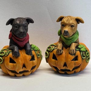 May include: Two ceramic figurines of dogs sitting inside orange jack-o'-lantern pumpkins. The dog on the left is black with a red bandana, and the dog on the right is brown with a green bandana.