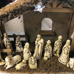 May include: A traditional nativity scene with white painted figurines of Mary, Joseph, baby Jesus, the three wise men, shepherds, and animals. The figures are arranged in a wooden stable setting with a star on top.