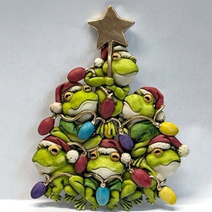 May include: A Christmas tree bauble featuring a stack of green frogs wearing Santa hats and holding colourful lights. The frogs are arranged in a pyramid shape with a gold star on top.