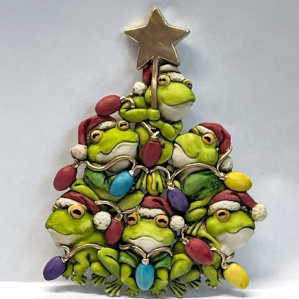 May include: A Christmas tree ornament featuring a stack of green frogs wearing Santa hats and holding colorful lights. The frogs are arranged in a pyramid shape with a gold star on top.