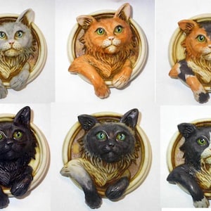 以下が含まれることがあります： 円形のフレームの中に、それぞれ異なる色と模様のセラミック製の猫の置物が6体飾られています。猫たちはすべてカメラを見ており、手足をフレームの縁に置いています。