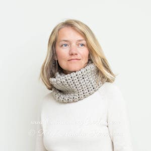 Pode incluir: Uma mulher usando uma echarpe de pescoço de gola alta de crochê bege. A echarpe é feita com um padrão de ponto texturizado. A mulher está usando um suéter branco e tem cabelo loiro.