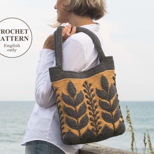 Può includere: Una borsa tote all'uncinetto con un motivo a foglie grigio e beige. La borsa ha una parte superiore e manici grigi, con un corpo beige con un motivo a foglie ripetuto. Il testo "CROCHET PATTERN English only" è in un cerchio.