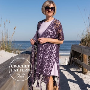 May include: A woman wearing a dark purple crochet shawl with a floral pattern and fringe, standing on a wooden boardwalk near the beach. The shawl is open, revealing a light purple dress underneath. The image includes the text "CROCHET PATTERN English Dutch".