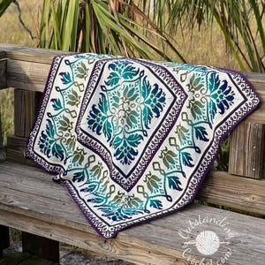 Può includere: Una coperta quadrata all'uncinetto con un motivo centrale a rombi e un bordo con intricati disegni floreali in sfumature di verde acqua, verde e viola scuro su sfondo bianco. Il testo OutstandingCrochet.com è visibile.
