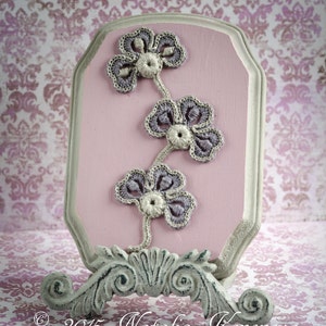 May include: A pink wooden plaque with a grey crocheted flower design. The plaque is on a white stand with a decorative scroll design. &copy; 2015 Natalia Kononova Outstanding Crochet.com