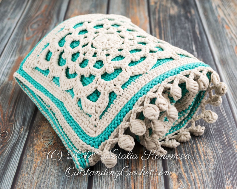 Puede incluir: Un coj&iacute;n de punto de crochet con un dise&ntilde;o de mandala blanco y turquesa. El coj&iacute;n tiene un borde de flecos con borlas.