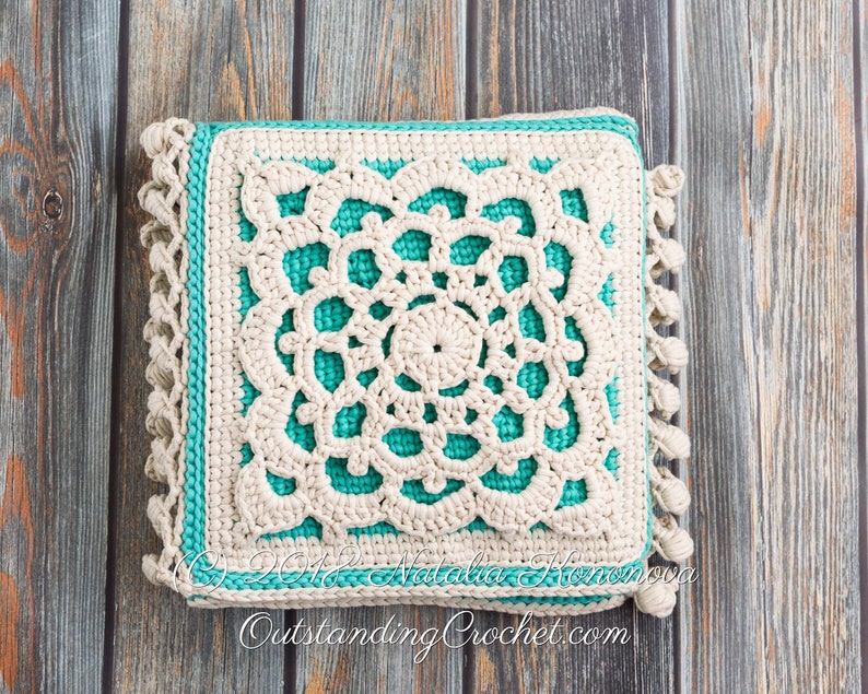 Puede incluir: Una almohada cuadrada de crochet con un dise&ntilde;o de crochet blanco sobre un fondo turquesa. La almohada tiene un borde de flecos y est&aacute; fotografiada sobre una superficie de madera.
