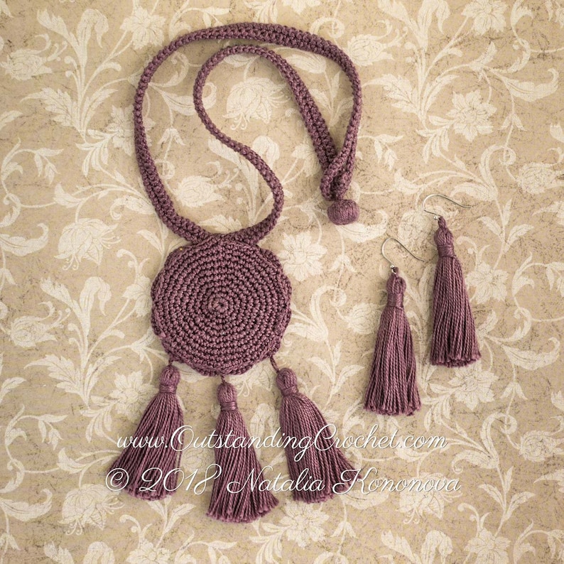 May include: A set of crocheted jewelry in a mauve color. The set includes a necklace with a round pendant and three tassels, and a pair of earrings with tassels.