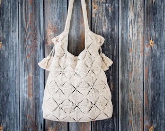 May include: A cream-colored crochet tote bag featuring a repeating diamond pattern throughout the body. The bag includes two long shoulder straps and two decorative tassels attached to the sides near the top opening of the bag.