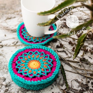 May include: Two crocheted coasters with a colorful mandala design. The coasters are teal, pink, orange, and yellow. A white mug is sitting on one of the coasters.