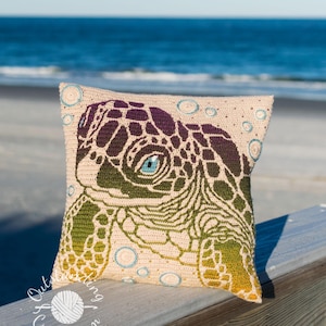May include: Crocheted cushion featuring a sea turtle design. The cushion has a beige background with a detailed turtle face in shades of green, purple, and yellow. The turtle has a blue eye. The cushion is decorated with blue circles.