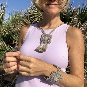 May include: A woman wearing a light purple sleeveless top with a crocheted necklace and bracelet. The necklace has a square pendant with a floral design and tassels. The bracelet is a wide band with a floral design.