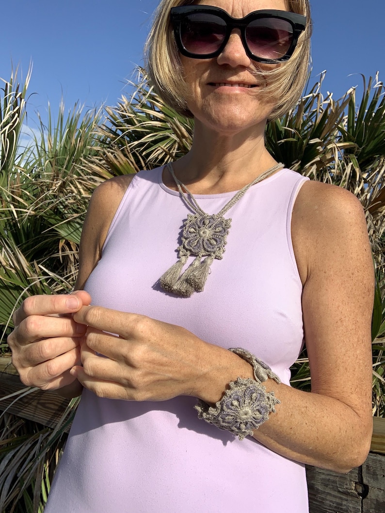 May include: A woman wearing a light purple sleeveless top with a crocheted necklace and bracelet. The necklace has a square pendant with a floral design and tassels. The bracelet is a wide band with a floral design.